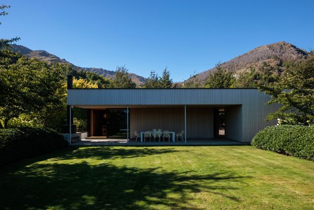 A low, single-storey modern house set within a manicured garden, with a long corrugated metal-clad roof and facade, extensive dark-framed glazing, and a deep covered outdoor area with dining furniture. Mountainous terrain rises behind the property under a clear blue sky.