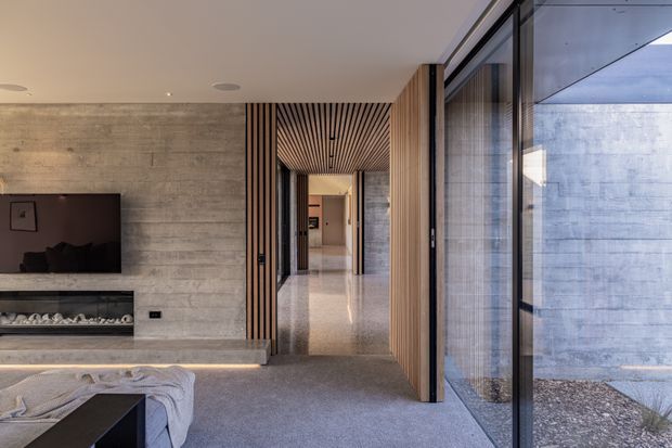 A refined contemporary living room opening onto a hallway, with a linear gas fireplace and wall-mounted television set into a raw concrete feature wall. Vertical timber battens frame the passage and continue across the ceiling, while full-height black-framed glazing overlooks a small enclosed courtyard. The room has a neutral palette, polished light flooring in the hall, and a large grey upholstered seating area in the foreground.
