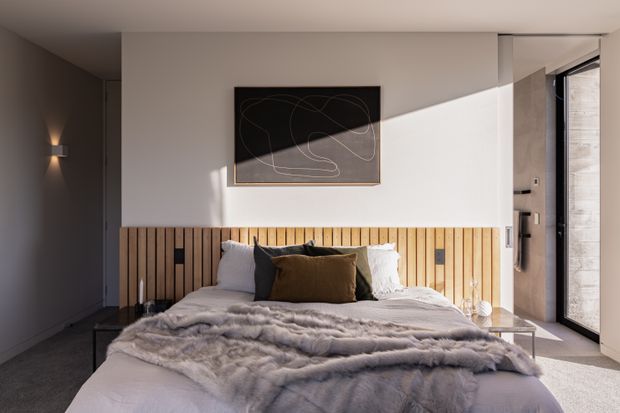 A contemporary bedroom with a low-profile bed dressed in layered grey bedding and pillows, set against a full-width vertical timber slat headboard. The room has smooth white walls, a dark grey side wall, a minimalist black artwork, concealed or simple lighting, and a large glazed door or window bringing in strong natural light. The palette is restrained and warm, with a refined, calming atmosphere.