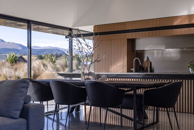 A contemporary open-plan kitchen and dining area with a long dark island and integrated dining counter, surrounded by floor-to-ceiling glazing overlooking a mountainous landscape. Warm timber cabinetry and vertical timber screening contrast with dark stone or composite surfaces, black-framed glazing, sculptural pendant lighting, and dark upholstered dining chairs. A sofa edge is visible in the foreground, suggesting an adjoining living area.
