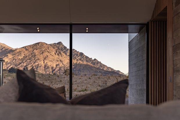 Interior view from a bedroom toward dramatic rugged mountains through expansive floor-to-ceiling glazing. The foreground shows the edge of a bed with dark pillows, while the room features a minimalist contemporary shell, dark metal glazing frames, a grey masonry wall, and vertical timber slatted detailing.