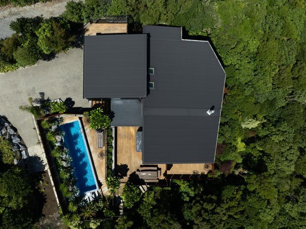 Aerial top-down view of a large dark-roofed house surrounded by dense greenery. Timber decking and outdoor entertaining areas are visible around a long rectangular swimming pool, with paved access and landscaped planting along the property edges.