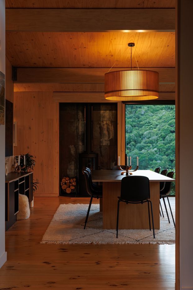 Warm, timber-lined dining room viewed through a doorway. The room features extensive honey-toned timber wall and ceiling lining, exposed dark timber beams, a large slatted pendant light, a rectangular dining table with black chairs, and a compact black wood-burning stove set against a dark feature wall. A large window frames dense green vegetation outside, while a textured neutral rug and simple shelving add softness to the space.