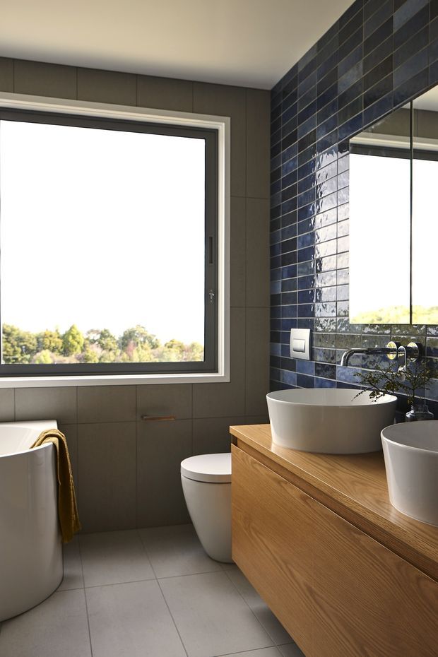 A bright contemporary bathroom with a freestanding white bathtub, toilet, double vessel basins on a timber vanity, large mirror, and generous window. Dark grey wall tiles and a glossy navy-blue tiled feature wall contrast with light grey floor tiles and warm timber cabinetry.