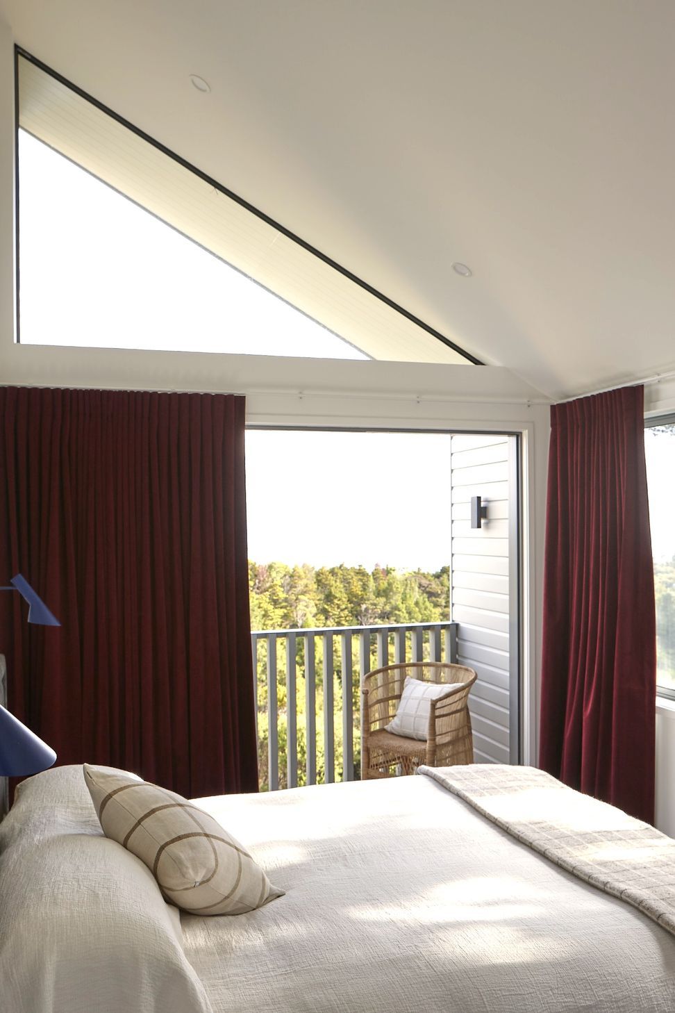 The main bedroom is a peaceful retreat featuring a soaring skillion ceiling, oversized glazing and direct access to a private deck. Maroon velvet curtains, blue AJ wall lights and a restrained material palette create warmth and character.