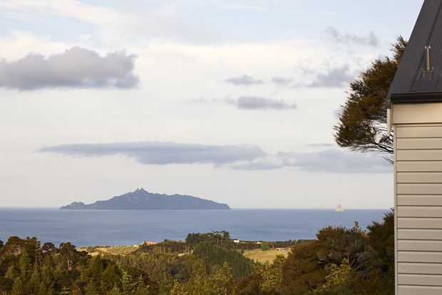 Exterior architectural view showing the side of a contemporary house with light horizontal weatherboard cladding and a steep dark roof edge, set above a coastal landscape overlooking the ocean and a prominent offshore island. The image is dominated by the building facade and expansive coastal setting.