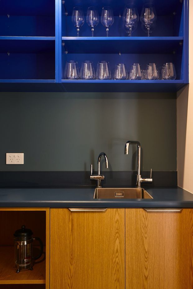 Close-up view of a compact, moody kitchen or bar joinery area. Deep navy-blue open shelving displays wine glasses above a dark benchtop and integrated stainless-steel sink with twin chrome mixer taps. Warm timber-grain cabinetry and open shelving occupy the lower section, with a French press in one cubby. The space has a dramatic, refined palette of navy, charcoal, chrome, and golden-brown timber.