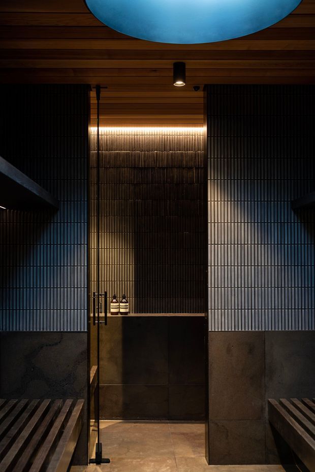 A dark, atmospheric sauna or steam-room interior with vertical black ribbed wall panels, warm timber ceiling boards, a central illuminated textured feature wall, dark stone or concrete lower walls, timber bench seating on both sides, and a glass door with black framing. A large blue circular ceiling light or skylight provides a cool contrasting accent.