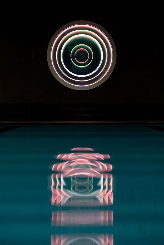 A dark, atmospheric view of a swimming pool reflecting a circular neon light installation mounted on a dark wall. The artwork consists of concentric glowing rings in pink, white, green, and blue tones, creating a vivid symmetrical reflection on the deep teal water. The scene is minimalist and highly dramatic, with the illuminated circle as the focal point.