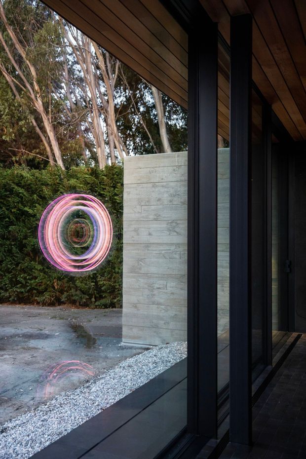 A contemporary architectural view from a dark, glazed interior or covered passage looking onto a landscaped courtyard. Full-height black-framed glazing and structural mullions line the right side, with a warm timber-lined soffit overhead. A pale horizontal-board concrete wall and white gravel strip border the courtyard, backed by dense trees and planting. A circular pink light-art installation is visible among the greenery, with its reflection appearing in the glass and on the wet paving.