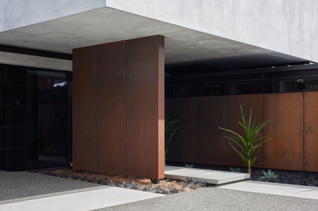 Contemporary residential exterior with dark timber or rust-toned metal cladding, a broad flat concrete canopy, black-framed glazing, and a minimalist landscaped entry path. Linear planters and sparse architectural planting reinforce the restrained modern composition.