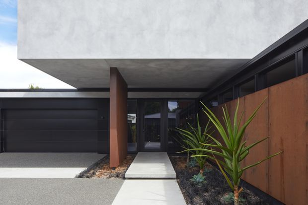 Modern residential entrance with a strong cantilevered concrete upper volume, dark garage door, recessed glass entry, rust-coloured structural column, and a landscaped path bordered by a corten-like metal screen. The composition is minimalist and geometric, with a restrained palette of concrete grey, charcoal, rust brown, and green planting.