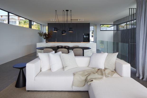 Contemporary open-plan living and dining area with a large white sectional sofa in the foreground, a dark kitchen island and cabinetry behind it, sculptural pendant lights, expansive glazing, and a vertical black screen feature near the windows. The restrained palette of white, charcoal, grey, and natural timber creates a clean, refined interior.