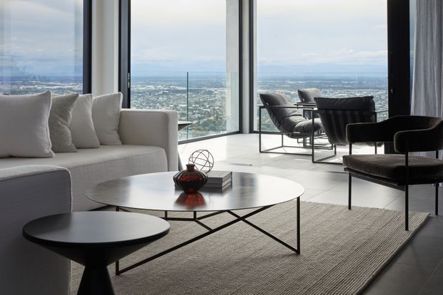 A sophisticated high-rise living room with floor-to-ceiling corner glazing overlooking a broad urban landscape. The room features a pale sectional sofa, two low round coffee tables, sculptural dark occasional chairs, a large neutral textured rug, and a minimalist black-framed dining or lounge setting near the windows. The bright, open interior has a refined contemporary aesthetic with strong geometric lines and expansive views.