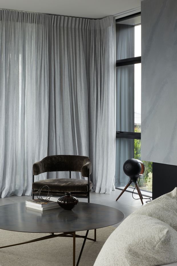 A refined contemporary living room with full-height sheer grey curtains, dark-framed floor-to-ceiling glazing, a sculptural brown armchair, a large round dark coffee table, and a light textured sofa. The room has a restrained neutral palette with black and dark brown accents, polished concrete or stone flooring, and minimalist decorative objects.