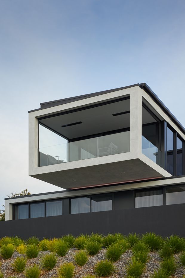 Exterior architectural photograph of a contemporary multi-level residence with a bold cantilevered upper volume, flat rooflines, dark cladding, pale concrete or rendered framing, and extensive floor-to-ceiling glazing. A covered balcony or living space projects over the lower level, with minimalist recessed ceiling lights visible beneath the overhang. The foreground contains designed gravel landscaping with ornamental grasses.