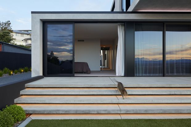 Exterior view of a contemporary multi-level residence with a low-profile flat roof, crisp white rendered walls, dark-framed floor-to-ceiling glazing, and a broad concrete terrace approached by wide stepped platforms. A bedroom is visible through the open sliding door, while the glazing reflects the evening sky and surrounding landscape.