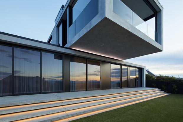 Contemporary multi-level residential exterior with extensive floor-to-ceiling glazing, dark framed windows, exposed concrete surfaces, and a dramatic cantilevered upper volume. Broad illuminated concrete steps lead from the glazed ground floor to a manicured lawn, with warm sunset reflections visible in the glass.