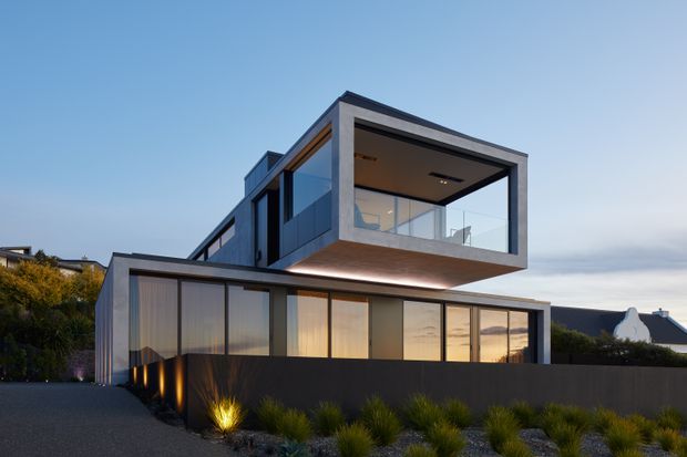 Contemporary multi-level residential house photographed at dusk, featuring bold cantilevered rectangular volumes, extensive floor-to-ceiling glazing, dark grey rendered walls, and a low front boundary wall. The landscaped frontage includes ornamental grasses and subtle exterior lighting, while warm interior light contrasts with the cool evening sky.