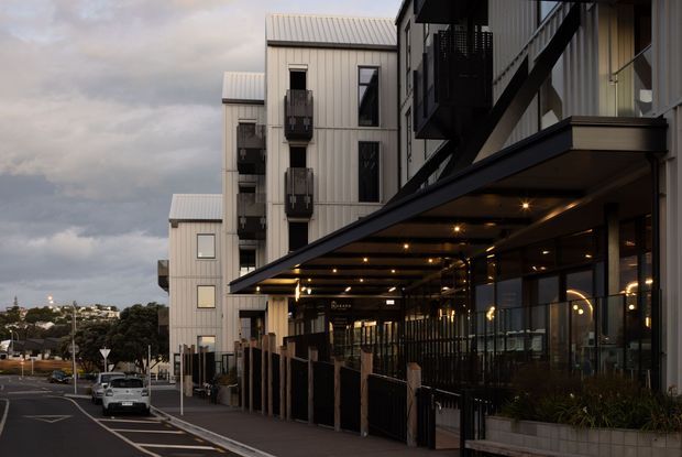 Exterior street-level view of a contemporary multi-storey building, likely a mixed-use or commercial development. The facade uses light vertical metal cladding, dark balcony and structural elements, extensive glazing, and a deep illuminated canopy over a covered pedestrian frontage. Urban streetscape and neighbouring buildings are visible in the background.