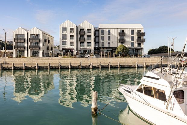 A waterfront multi-storey residential development viewed across a marina. Several matching apartment buildings have pale vertical cladding, steep gabled roof forms, dark-framed windows, and projecting balconies. Boats and calm water occupy the foreground, with a clear blue sky behind the development.