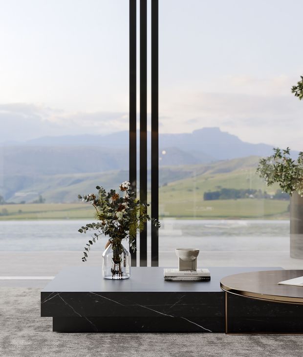 A refined contemporary interior vignette centered on a long black marble console with a rounded adjoining table. A sculptural floral arrangement, books, and a small vessel sit on the console, while full-height glazing with slender black mullions frames a serene lake and mountain landscape beyond. The room uses a restrained palette of black, grey, white, and natural tones.