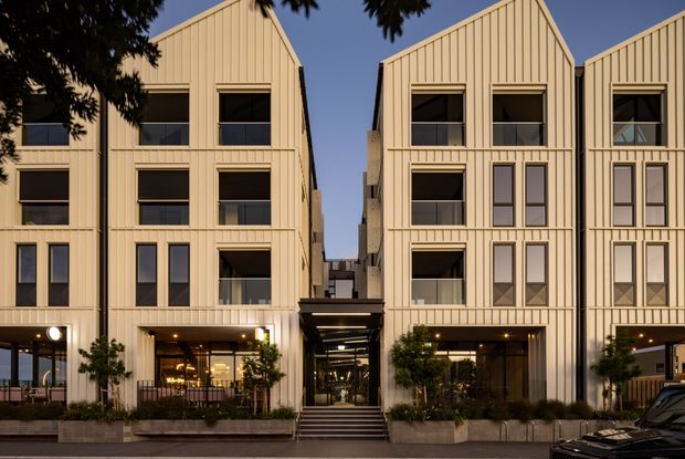 Street-level exterior view of a contemporary multi-storey residential development composed of several narrow gabled buildings. The façades use pale vertical cladding with regularly spaced windows, recessed balconies, ground-floor glazed commercial or communal spaces, and a covered pedestrian entrance between the buildings. Planter boxes and small trees line the frontage.