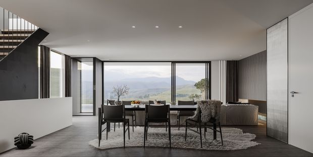 A refined, open-plan dining and living area with a contemporary minimalist interior. A dark dining table and black chairs sit on a textured rug beside a low neutral sofa, with expansive black-framed glazing overlooking a mountainous landscape. A stairway rises at the left, while a tall flush internal door and vertical wall detailing add architectural interest.
