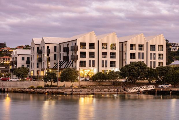 A dusk waterfront view of a contemporary multi-storey residential or mixed-use building with repeated steep gabled roof forms, pale vertical cladding, extensive dark-framed windows, balconies, and landscaped trees along the waterfront promenade. Reflections of the illuminated building appear in the calm water in the foreground.