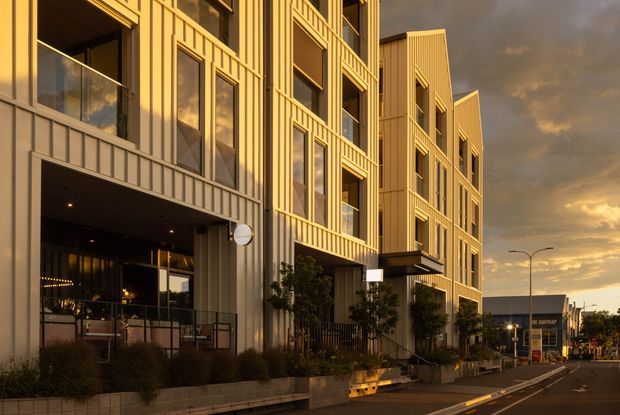 Exterior street-level view of a contemporary multi-storey urban building with a strong vertical facade rhythm, projecting window bays, recessed balconies, and pale metal-clad surfaces. Ground-floor commercial or lobby glazing opens onto a landscaped frontage, while the building extends along a marked street toward other urban buildings. Warm low-angle sunlight and dark clouds create a dramatic golden-hour atmosphere.