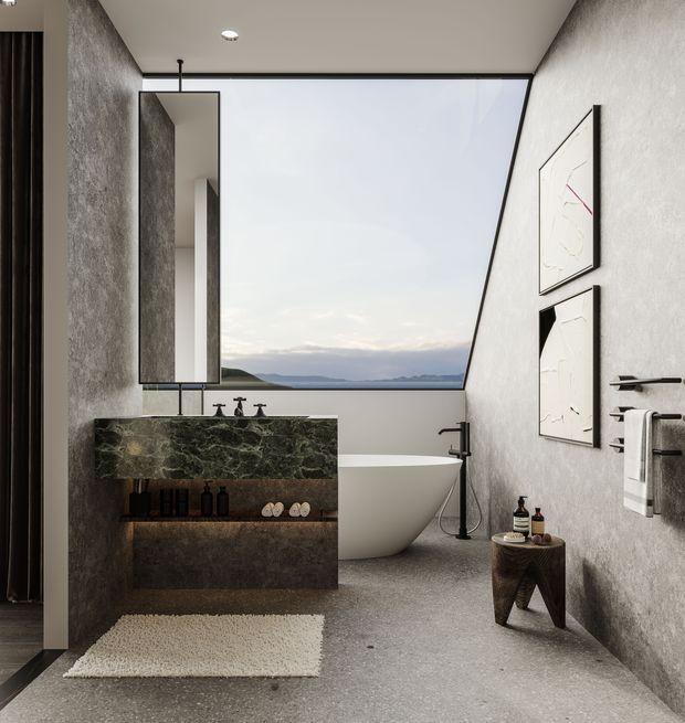 A refined contemporary bathroom with textured grey walls and flooring, a freestanding white bathtub positioned beside expansive angled glazing, and a dark green veined stone vanity with an integrated basin. A tall frameless mirror, black tapware, wall-mounted towel rails, minimalist artwork, and small timber side table complete the sophisticated interior. A distant landscape is visible through the window.
