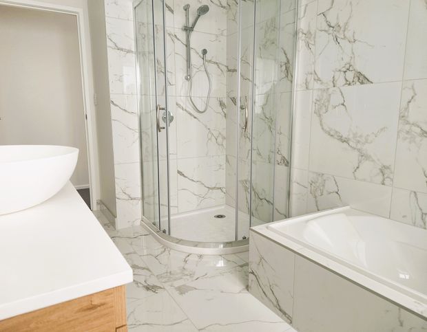 Bright contemporary bathroom featuring a glass-enclosed corner shower, white freestanding basin on a timber vanity, and a white bathtub. Large white marble-look tiles with prominent grey veining cover the walls and floor, complemented by chrome shower and tapware.