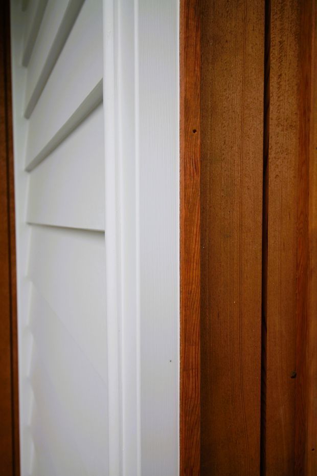 Close-up architectural detail showing white horizontal weatherboard-style cladding beside a vertical warm-toned timber wall or door, separated by a white trim. The image emphasizes contrasting painted and natural timber surfaces, with no broader room context visible.