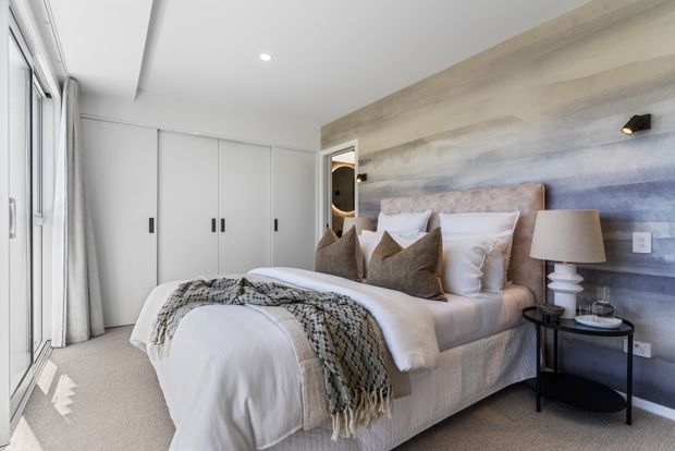 A bright, contemporary bedroom with a neatly made upholstered bed, layered neutral pillows, a textured throw, full-height white wardrobe cabinetry, and a feature wall combining pale timber-look horizontal panels with a softly blue-grey textured surface. Floor-to-ceiling glazing with sheer curtains provides abundant natural light, while a small black side table and sculptural table lamp sit beside the bed.