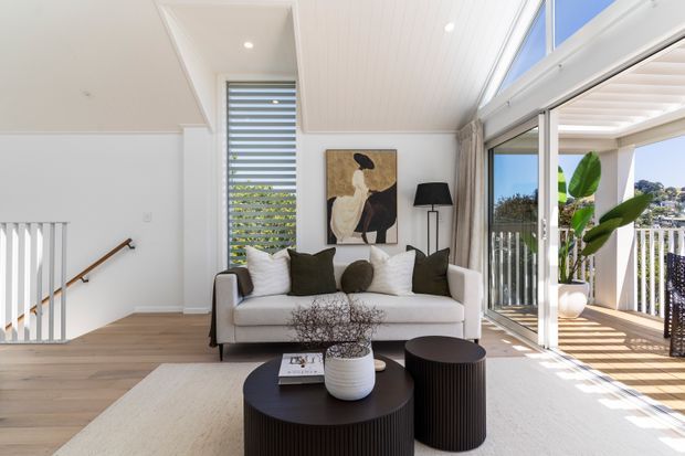 Bright contemporary living room with a white sofa, dark round coffee tables, neutral rug, abstract artwork, and a black floor lamp. The room has light timber flooring, white vertical wall and ceiling panelling, a stairwell with timber handrail, and extensive sliding glass doors opening to a covered balcony or terrace. Strong daylight, high clerestory glazing, and indoor-outdoor connection define the space.