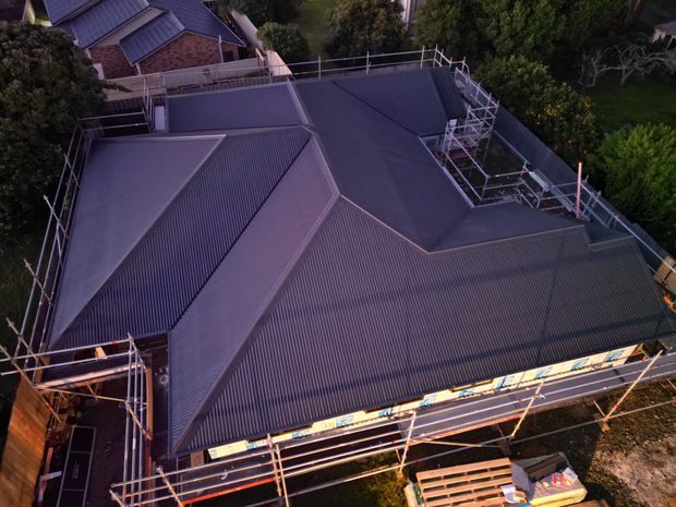 Aerial view of a large residential roof under construction or renovation, with dark corrugated metal roofing, multiple pitched roof sections, and extensive perimeter scaffolding. A neighbouring brick house and surrounding trees are visible in the background.