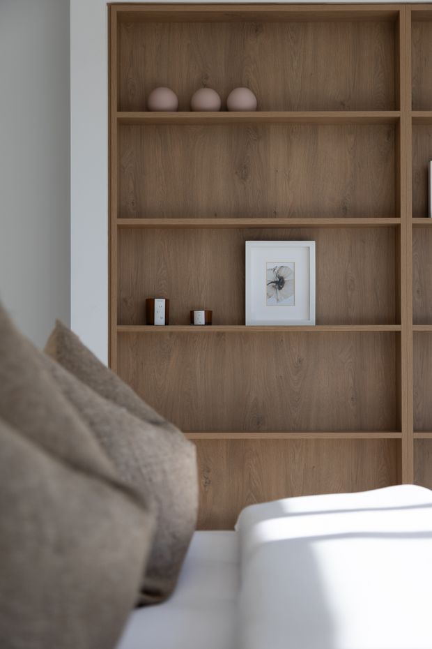 Close-up interior view of a warm timber built-in shelving unit with open compartments, displaying a few minimalist decorative objects and framed artwork. The edge of a neutral upholstered sofa and a white seating or furniture element are visible in the foreground. The composition is softly lit and restrained, with natural wood grain, pale walls, and a calm contemporary feel.