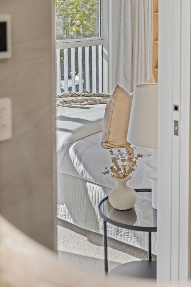 A softly lit bedroom viewed through a partially open doorway. The room features a white upholstered bed with layered white bedding, a textured beige accent pillow, a round black-framed side table with a cream ceramic vase and dried branches, a white table lamp, pale curtains, built-in timber shelving, and a large window overlooking a leafy exterior. The overall look is refined, neutral, and airy.