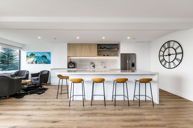 Bright open-plan kitchen and living area with a large white island breakfast bar, five timber-and-black bar stools, integrated white cabinetry, warm wood-grain overhead joinery, stainless-steel refrigerator, and pale timber-look flooring. A dark seating area and artwork are visible to the left, with a large black wall clock on the right. Recessed ceiling lighting and broad glazing create an airy contemporary interior.