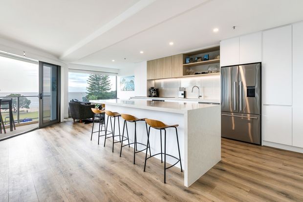 Bright open-plan kitchen and living area with a large white island, four timber-seated bar stools, integrated white cabinetry, stainless steel refrigerator, timber open shelving, and warm timber-look flooring. Full-height glazing and a sliding door open onto a balcony with coastal water views.