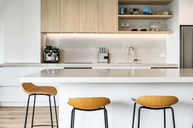 Bright contemporary kitchen featuring pale timber overhead cabinetry, open display shelving, a light stone island benchtop with integrated seating, a pale splashback, stainless-steel tapware and appliances, and three timber-and-black-metal bar stools. The space has a clean, refined and airy appearance.