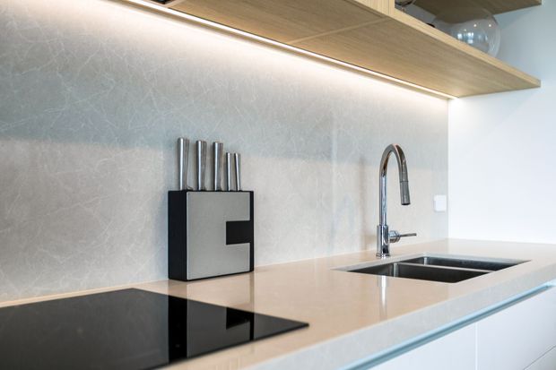 Close-up view of a contemporary kitchen benchtop featuring a pale stone surface, dark glass cooktop, undermount double sink, chrome gooseneck tap, and light timber overhead shelving with integrated under-shelf lighting. A pale textured splashback and white cabinetry create a clean, refined appearance.