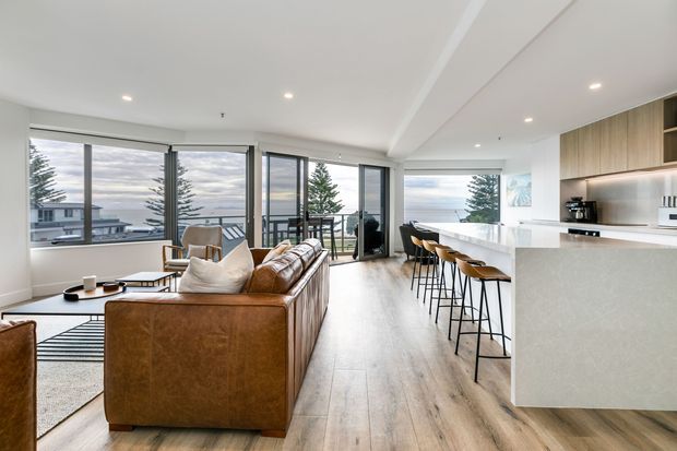 Open-plan contemporary living, dining and kitchen area with floor-to-ceiling glazing, a large leather sectional sofa, timber-look flooring, and a bright white kitchen island with bar seating. The space overlooks a coastal or suburban landscape and uses a restrained neutral palette with warm timber and leather accents.