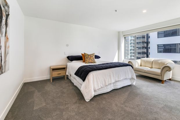 Bright, spacious contemporary bedroom with a large bed, neutral bedding, grey carpet, a simple timber bedside table, and a cream upholstered sofa positioned beside expansive apartment windows overlooking nearby high-rise buildings. White walls and ceiling create a clean, airy interior.
