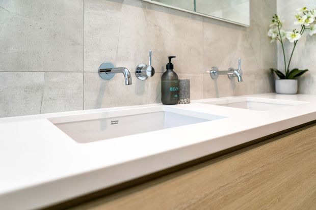 Close-up of a contemporary bathroom vanity with a wide white double basin countertop, wall-mounted chrome tapware, pale large-format tiled walls, and light timber cabinetry. A small orchid and soap dispenser provide subtle styling accents.