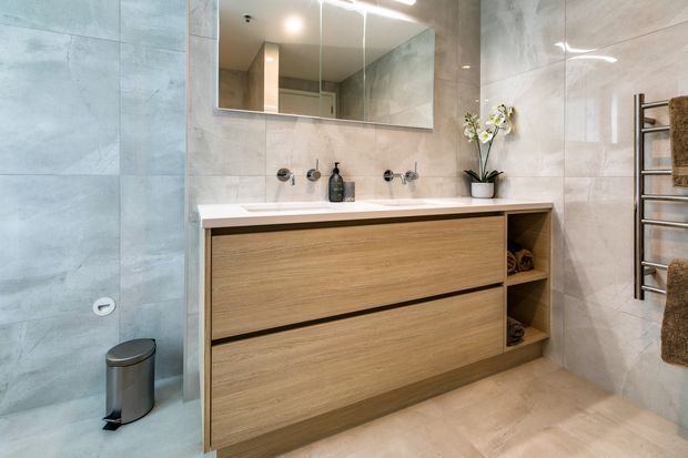 Contemporary bathroom with large pale stone-look wall and floor tiles, a floating timber-look double-drawer vanity, white benchtop basin, wall-mounted taps, mirrored cabinet, stainless-steel heated towel rail, and recessed shelving. The neutral, clean-lined fit-out is softly illuminated and includes a small orchid and folded towels.