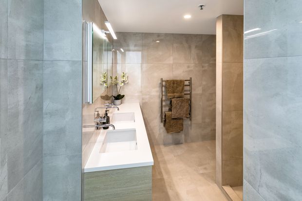 A contemporary bathroom viewed through a tiled opening, featuring a long white double vanity with integrated basins, wall-mounted mirrors, pale timber-toned cabinetry, large beige stone-look tiles, recessed ceiling lighting, and a towel ladder in the shower area. The space has a clean, warm-neutral and highly streamlined appearance.