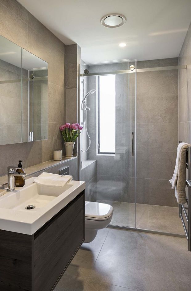 A compact contemporary bathroom with a wall-mounted dark wood vanity, white integrated basin, large mirrored cabinet, grey stone-look wall and floor tiles, and a frameless glass shower enclosure. A wall-hung toilet, recessed ceiling lights, narrow window, chrome fixtures, and a heated towel rail are visible. The neutral grey and white palette is softened by a small vase of pink tulips.
