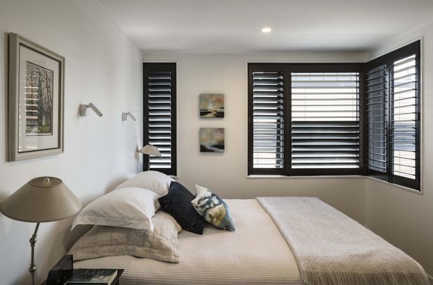 A calm, compact bedroom with a neatly made bed, layered neutral bedding, dark accent cushions, framed artwork, bedside lighting, and extensive dark plantation shutters across the windows. The room has white walls, recessed ceiling lighting, and a restrained contemporary interior.