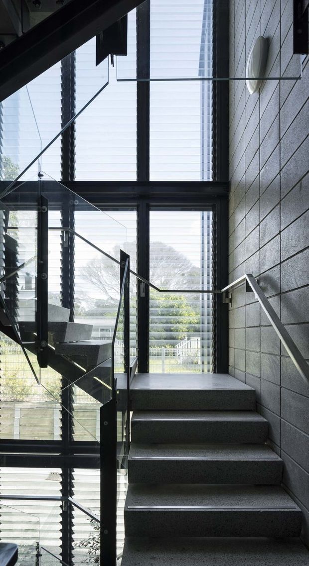 Interior view looking up a modern stairway with dark concrete or masonry finishes, a black structural frame, glass balustrades, and metal handrails. Large floor-to-ceiling windows with horizontal louvres provide filtered daylight and views of trees and neighbouring buildings. The stair rises alongside a tall glazed void, creating a dramatic, industrial-contemporary circulation space.